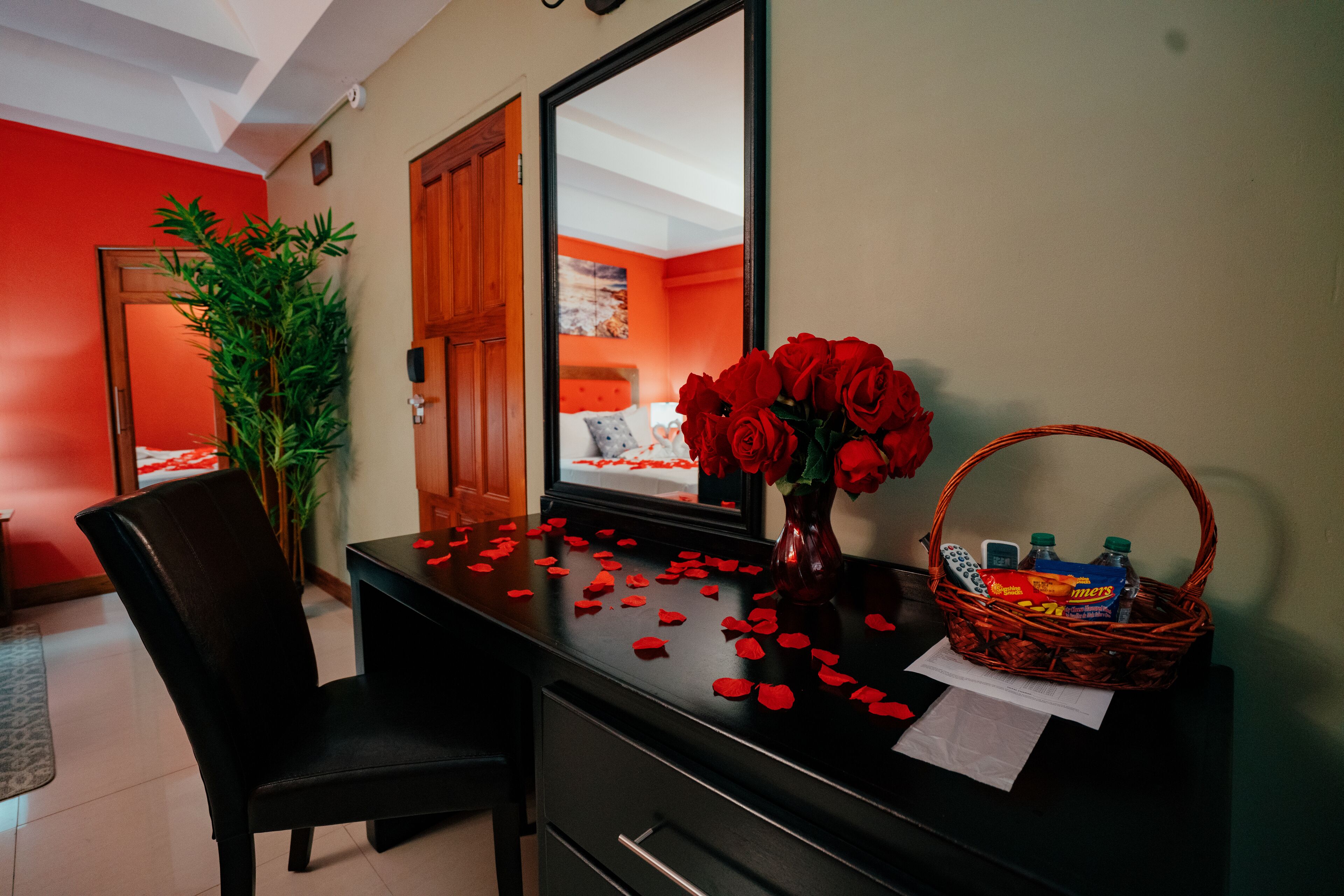 A hotel room with a desk, a chair, a mirror, a vase with red roses, a basket with snacks, and scattered rose petals.