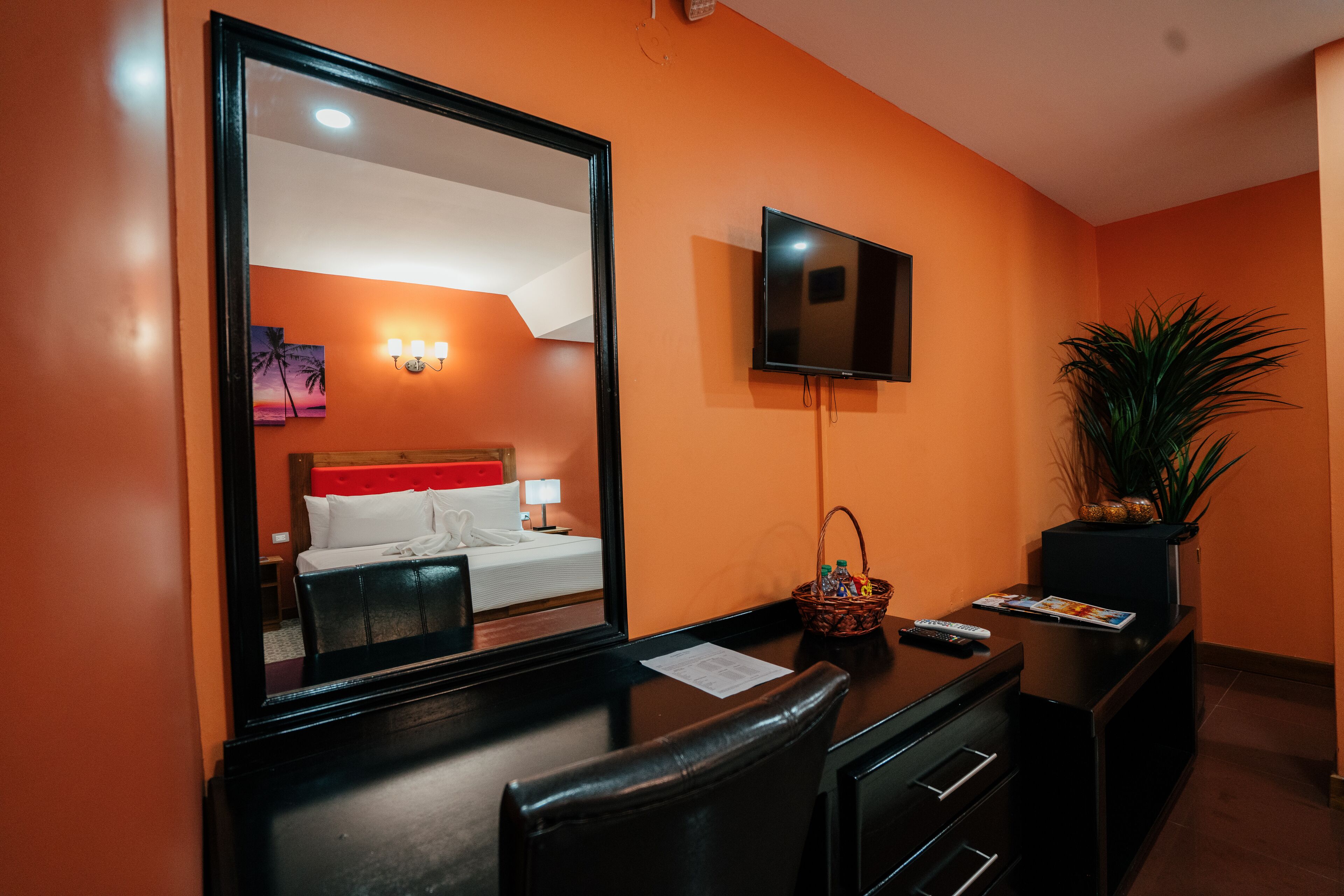 A hotel room with a large mirror, a flat-screen TV mounted on the wall, a desk with a basket, and a potted plant.