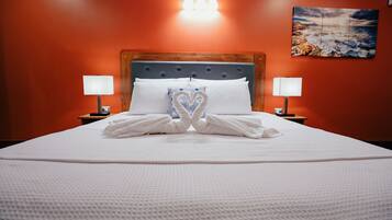 A neatly made bed with towels arranged in a heart shape, a wooden headboard, two bedside lamps, and a painting of a coastal scene on the wall.