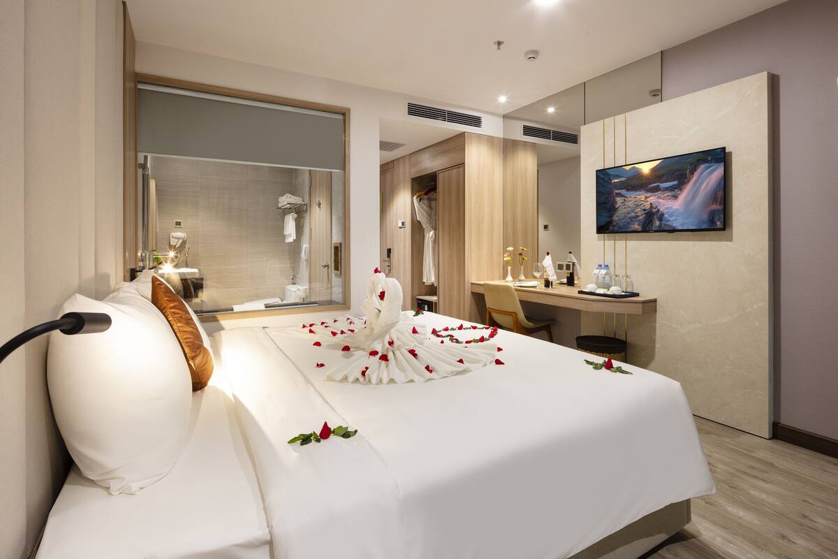Habitación de hotel con cama, escritorio, televisor y baño visible a través de una mampara de vidrio.
