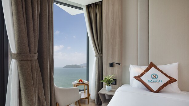 A hotel room with a bed, a nightstand, a small table with a plant, and a view of the sea. - Nalicas Hotel (Nha Trang)