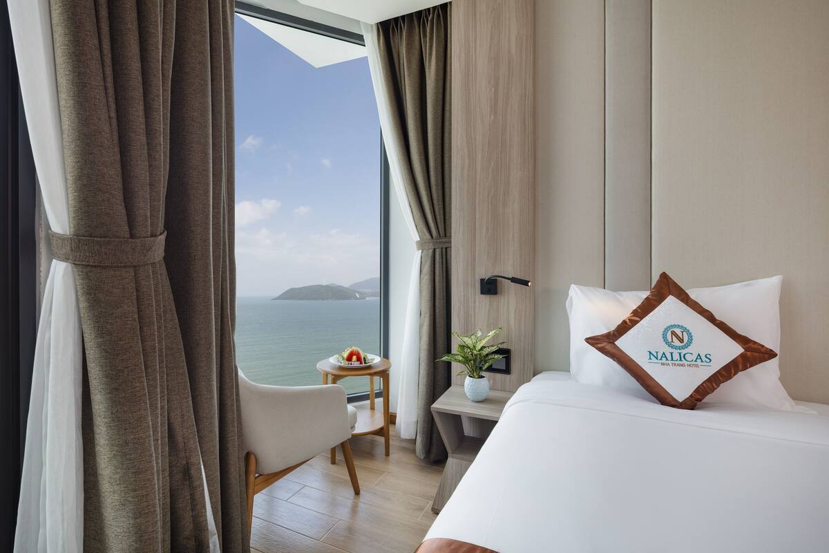 Habitación de hotel con cama, mesita de noche, pequeña mesa con planta y vista al mar.