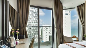A hotel room with a large window, a desk with a telephone, a chair, and a bed with a view of the city and sea. - Nalicas Hotel (Nha Trang)