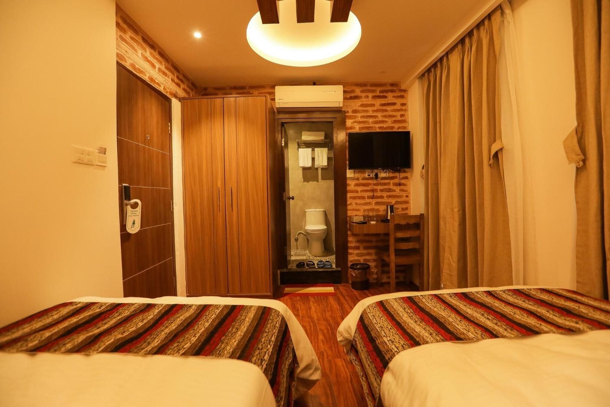 A hotel room with two beds, a television, a brick wall, and a bathroom visible through an open door.