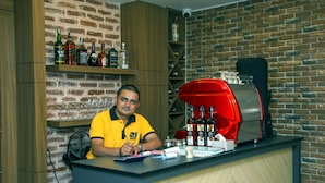A bar area with a brick wall, shelves with bottles, a red espresso machine, and a person in a yellow shirt. - Flying Yak Hotel - Hostel (Kathmandu)