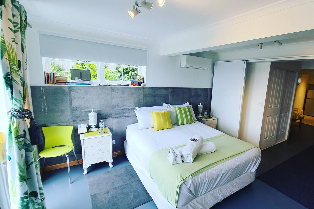 A modern hotel room with a large bed, a bedside table, a chair, and a window with a view of greenery.