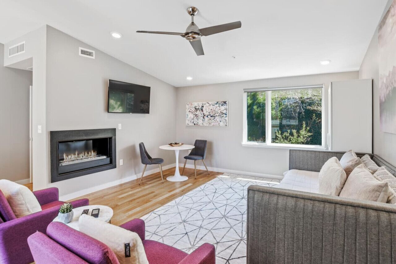 A modern living room with a fireplace, a ceiling fan, a large window, and a geometric rug.