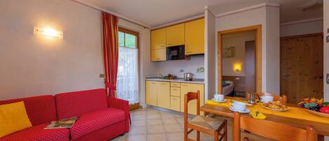 Un espacio habitable compacto con sofá rojo, mesa de comedor de madera, armarios amarillos y una zona de cocina.