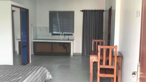 A small, modern room with a bed, a desk, a chair, a window with blinds, and an air conditioning unit. - MN Apartment (Siem Reap)