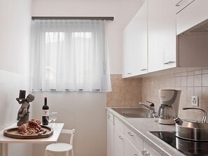 Une cuisine moderne avec des meubles blancs, une fenêtre ornée de rideaux légers, une machine à café et une bouilloire posées sur le plan de travail.