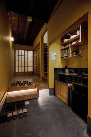 A traditional Japanese-style room with tatami flooring, a low wooden table, and a kitchen area with a sink and black tiled backsplash. - Tsumugi Horikawakichimonji (Kyoto)