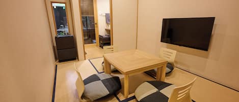 A small room with tatami flooring, a low wooden table, two chairs, a television, and a mirror.