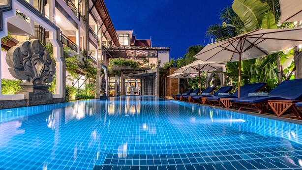 A swimming pool with tiled flooring, lounge chairs, umbrellas, and a building with balconies and plants. - Araya Angkor Residence (Siem Reap)