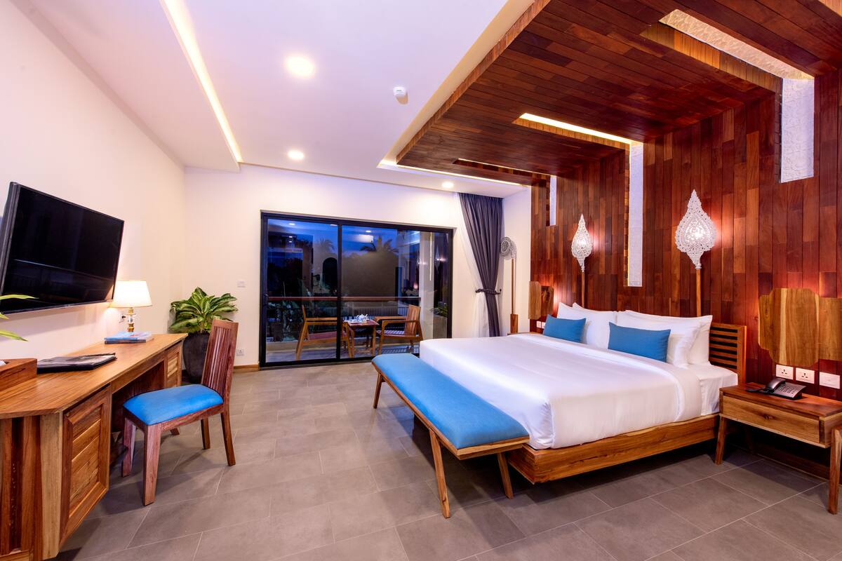 A modern hotel room with a large bed, a desk with a chair, a TV, and a balcony with a view.