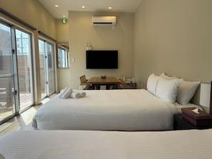 A hotel room with a large bed, a television mounted on the wall, a table with chairs, and a sliding glass door leading to an outdoor area. - Okinawa Green Lodge (Chatan)