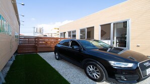 Un SUV Audi negro estacionado en un camino de acceso estrecho, con una cerca de madera y un edificio de fachada color crema.