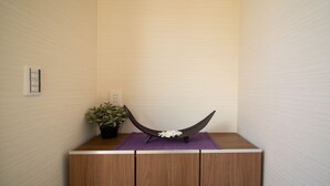 Una esquina de una habitación con un mueble de madera, un objeto negro y curvo sobre una alfombra morada, y una planta en maceta.