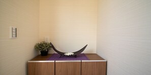 Una esquina de una habitación con un mueble de madera, un objeto negro y curvo sobre una alfombra morada, y una planta en maceta.