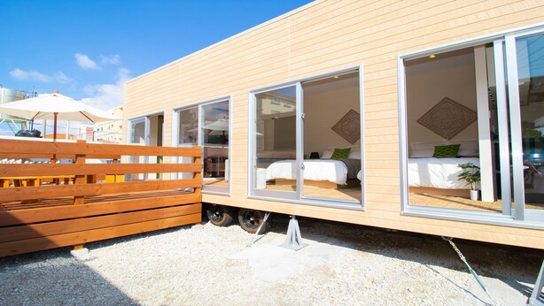 Un edificio moderno de un solo piso con ventanas amplias, una terraza de madera y un dormitorio visible con cama y almohadas verdes.