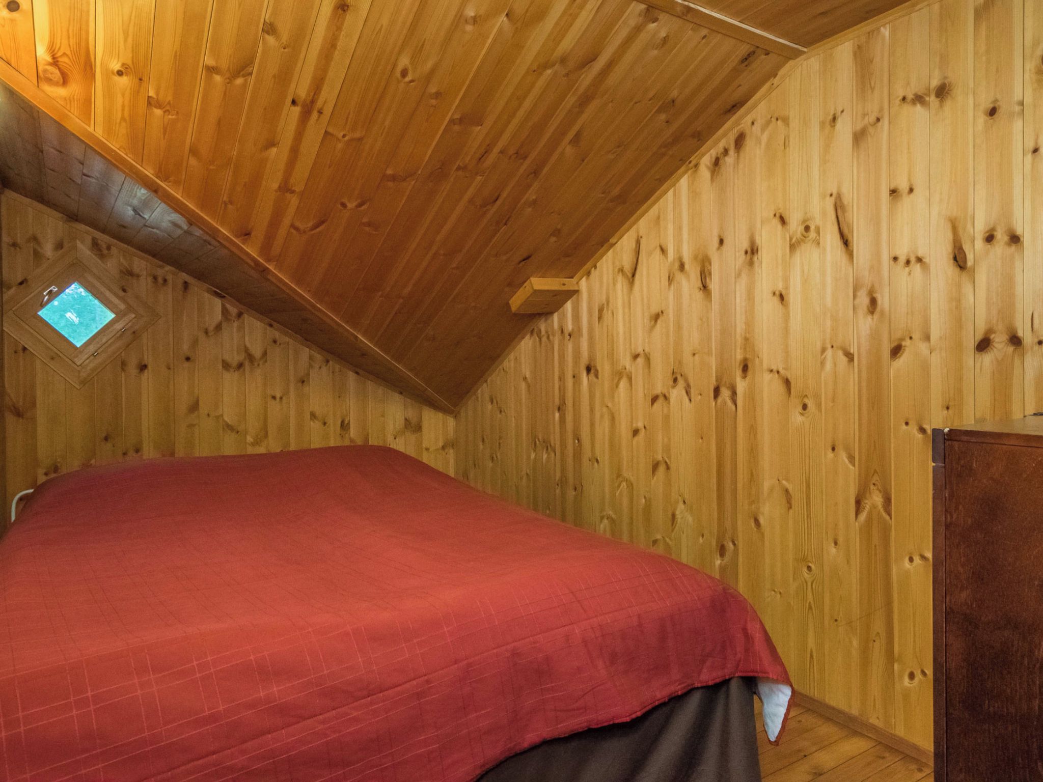 Una habitación con techo inclinado, paredes de madera, una cama con cubierta roja y un tragaluz pequeño.
