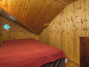 A room with a sloped ceiling, wooden walls, a bed with a red cover, and a small skylight.