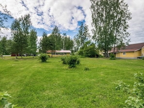 A grassy area with a building that has a red roof and a yellow exterior. There are trees and a clear sky with clouds.