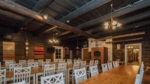 Un vaste espace dédié à la salle à manger, avec de longues tables en bois et des chaises blanches, une cheminée en briques et un plafond en bois.