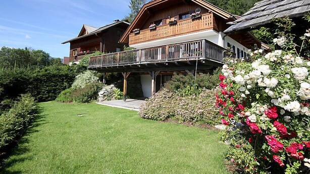 Eine zweistöckige Holzhütte mit Balkon, umgeben von einem gepflegten Garten und einem Wald im Hintergrund.