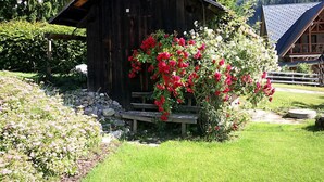 Eine rustikale Holzhütte mit geneigtem Dach, umgeben von üppigem Grün und einem blühenden Strauch.