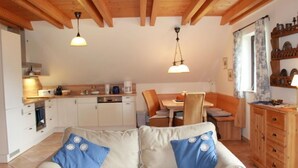 Ein gemütliches Wohnzimmer mit einer Couch und einem Esstisch, Holzbalken an der Decke und einer Küche im Hintergrund.