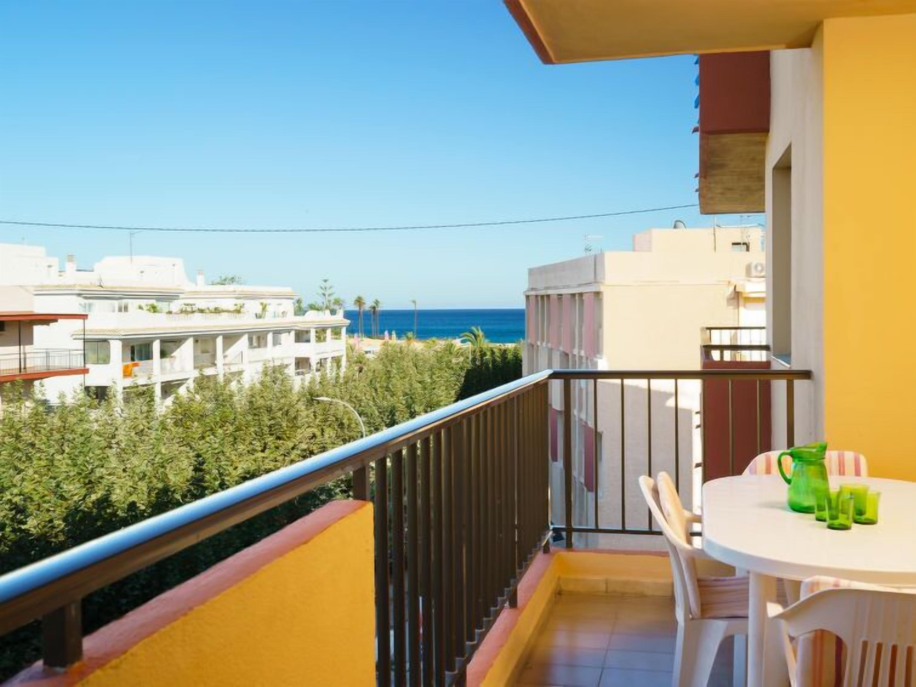 A balcony with a white table and chairs, a green pitcher, and a view of the sea and neighboring buildings.