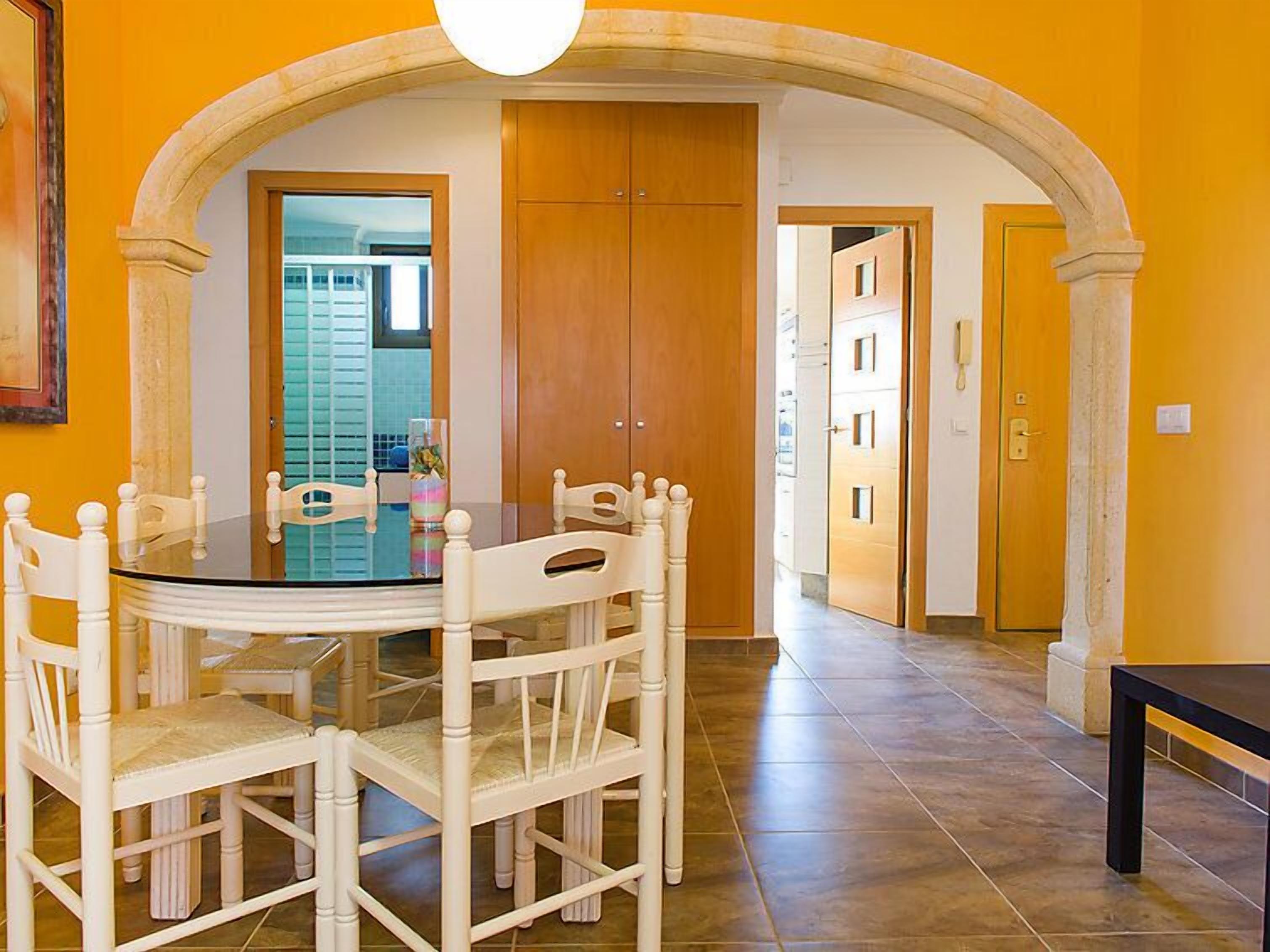 Un comedor con una mesa de vidrio rodeada de sillas blancas, un arco y puertas de madera.