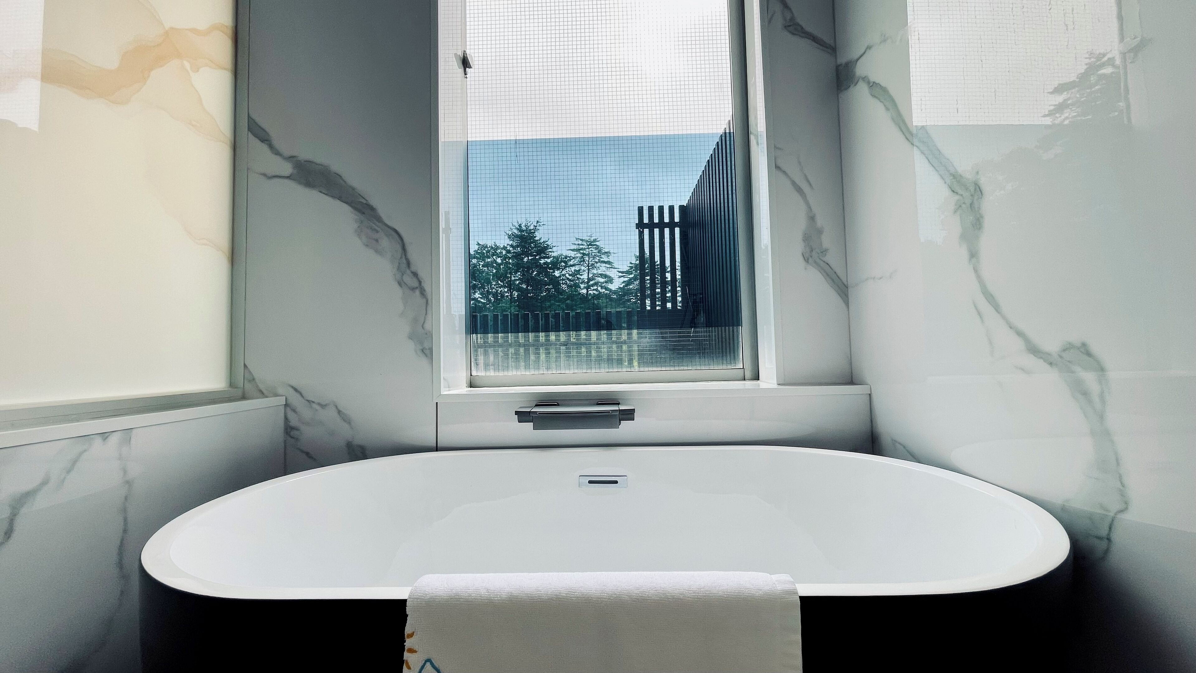 A modern bathroom with a freestanding bathtub, a window with a view of trees, and marble walls.