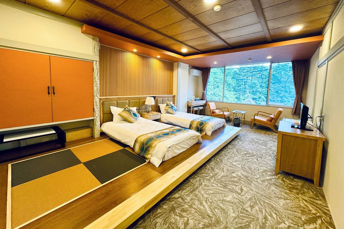 A hotel room with two beds, a desk, a chair, and a view of trees through the window.