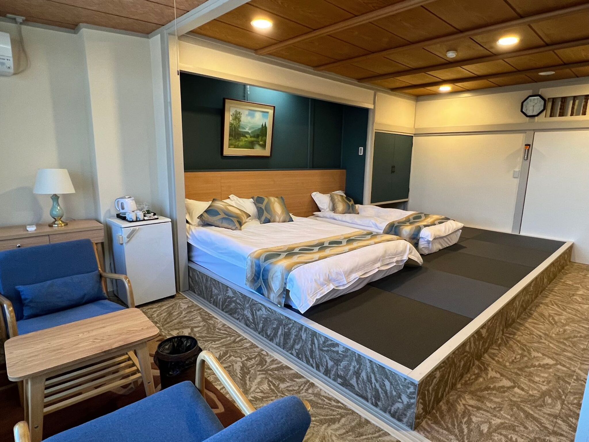 Ein Hotelzimmer mit zwei Betten, einem Schreibtisch, einem Stuhl, einem kleinen Tisch und einer Wanduhr.