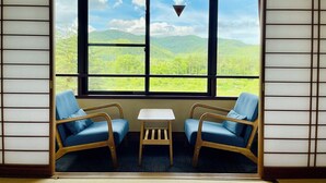 A room with a large window offering a view of green hills and a clear sky. There are two blue chairs and a small table in the room. - Kurobe Sun Valley Hotel (Omachi)