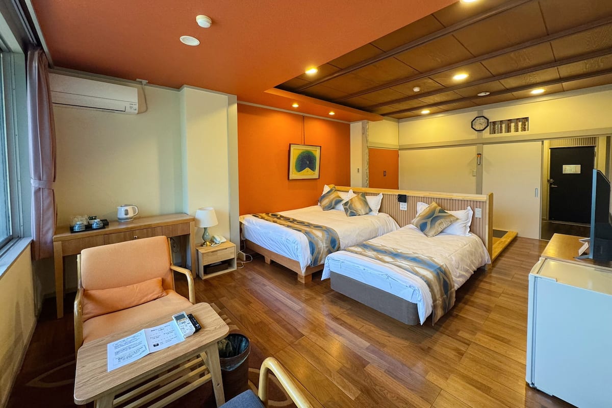 A hotel room with two beds, a wooden desk, a chair, a small table with a lamp, and a trash bin.