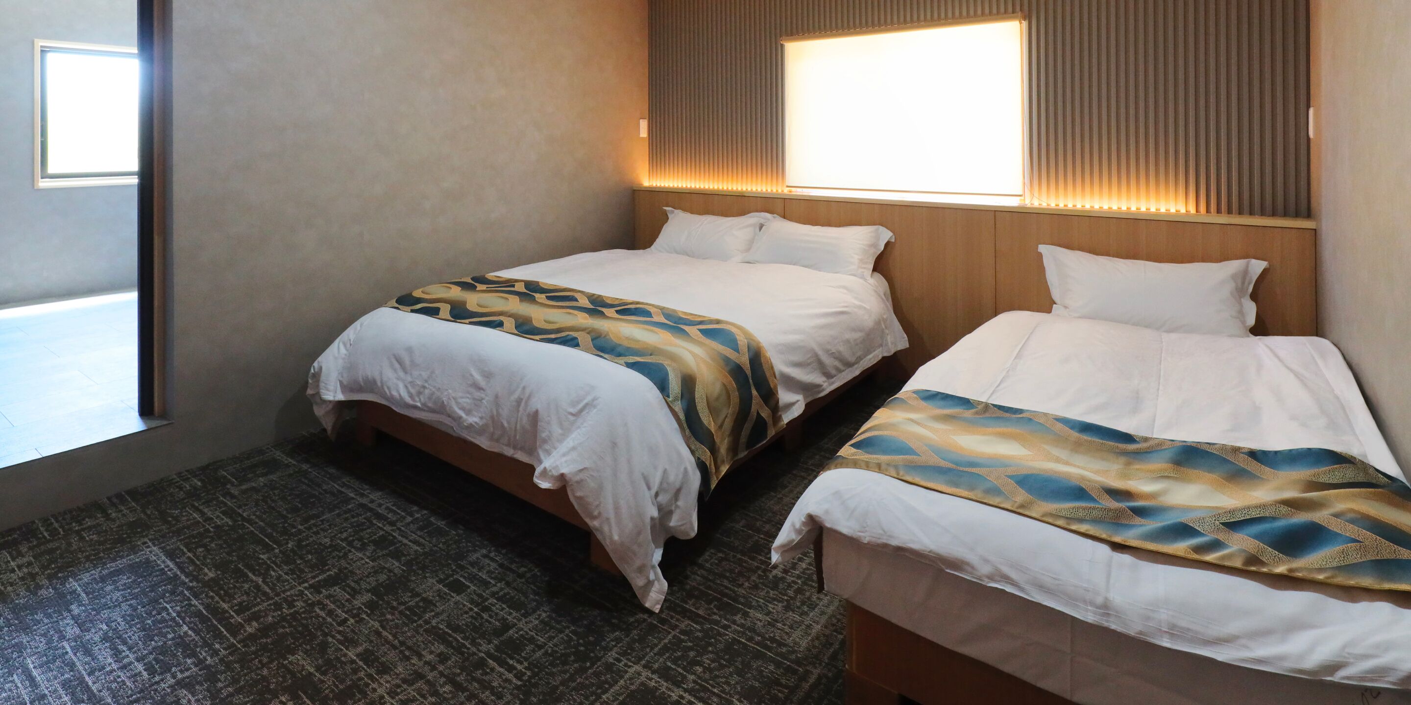 A hotel room with two beds, a patterned bedspread, a wooden headboard, and a window with blinds.