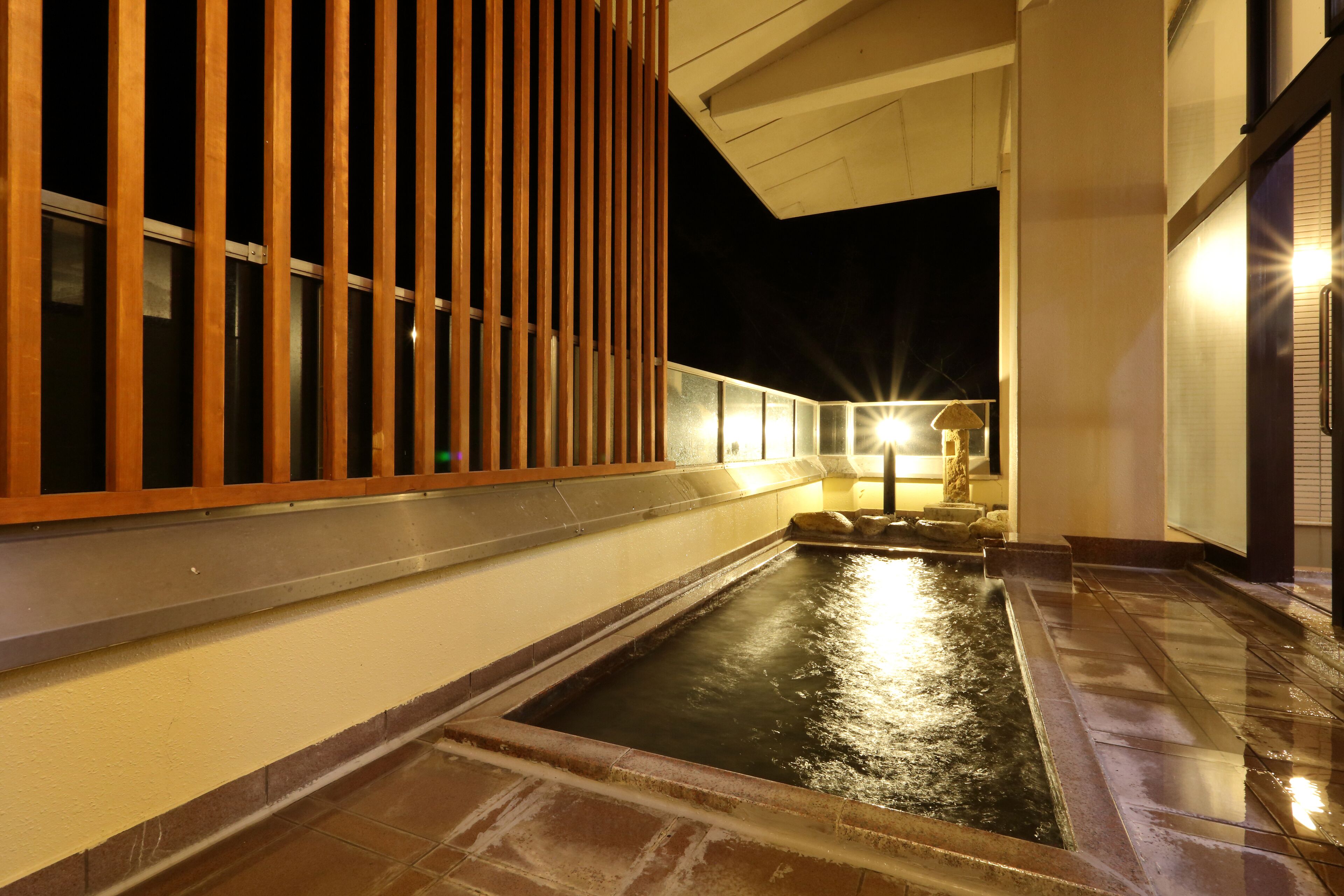 A nighttime view of a courtyard with a rectangular water feature, wooden slats, and a stone pathway.