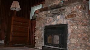 Ein Steinkamin mit hölzernem Sims, eine Kommode aus Holz mit zahlreichen Schubladen und eine Lampe auf einem Tisch.
