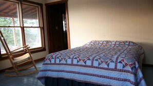 Ein Schlafzimmer mit einem Bett, das mit einer gemusterten Steude bezogen ist, einem Schaukelstuhl und einem Fenster mit Blick auf Bäume.