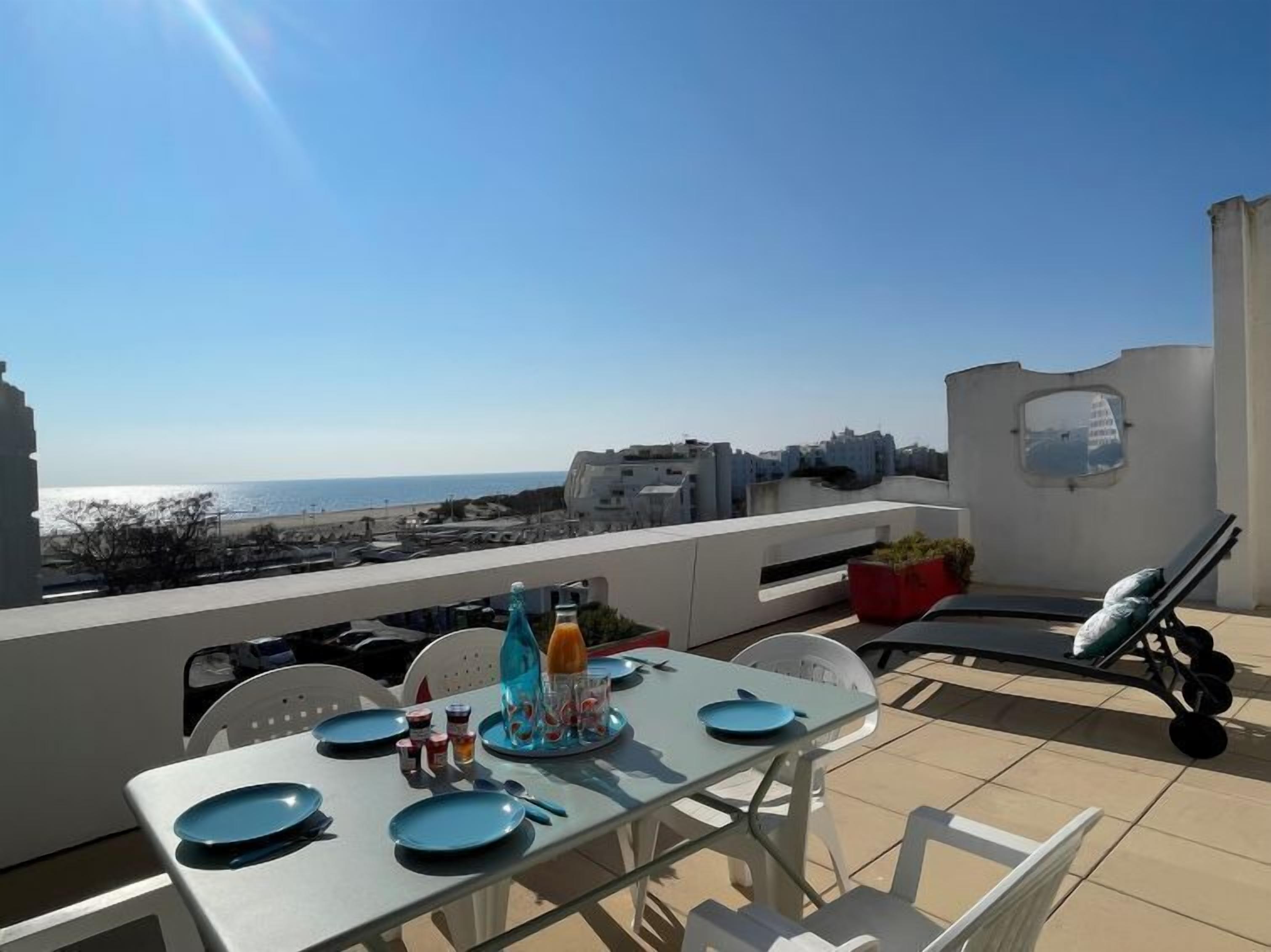 A rooftop terrace with a table set for two, overlooking a beach and cityscape.