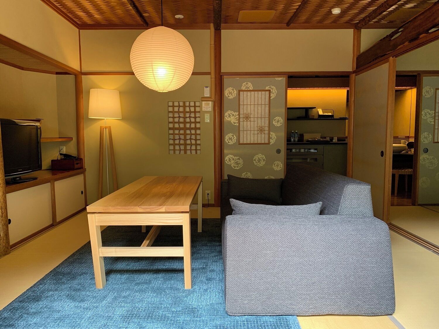 A traditional Japanese room with tatami flooring, a low wooden table, a sofa, a television, and a hanging paper lantern.