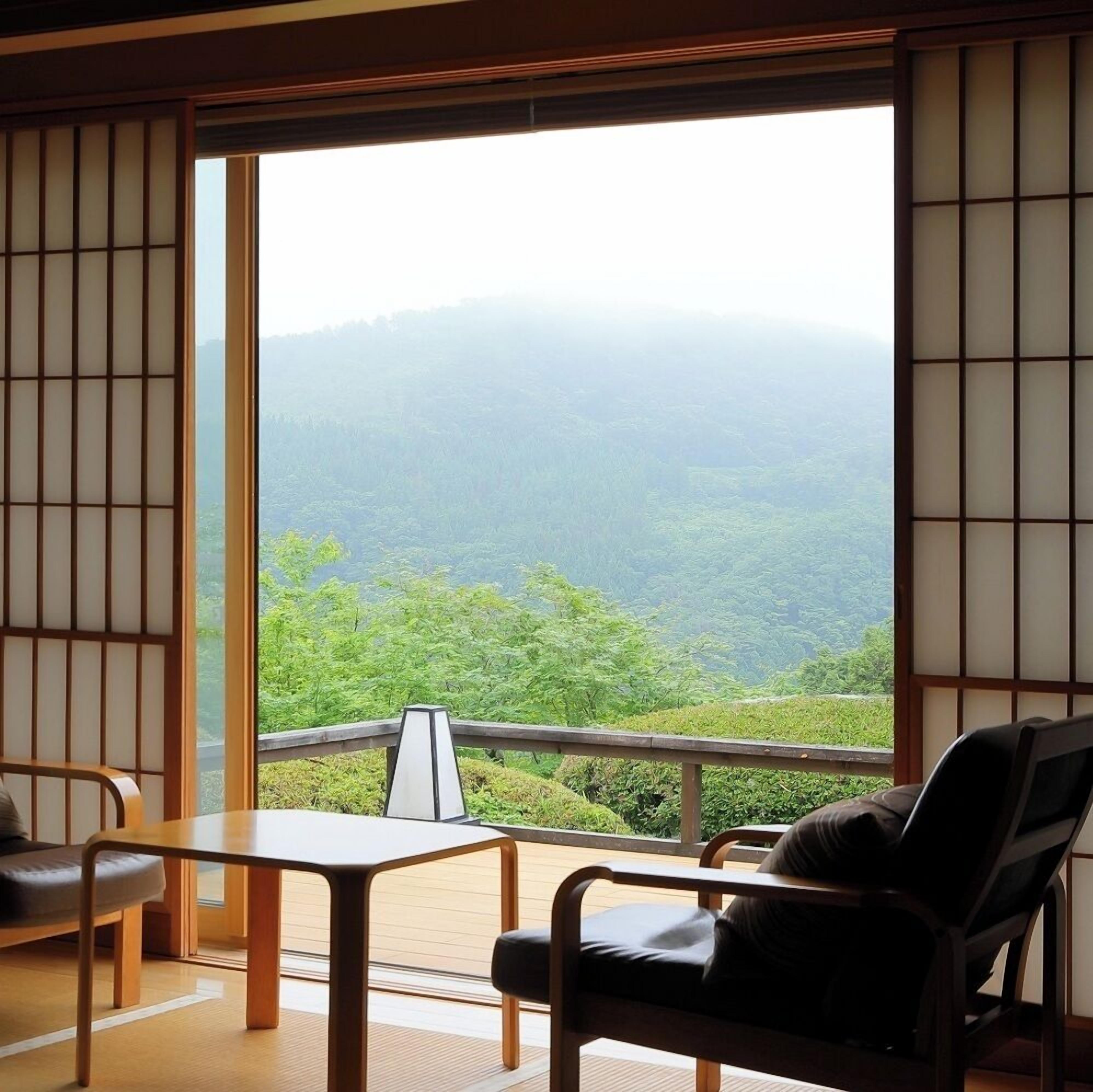 A room with tatami flooring, a low table, a chair, and a view of a forested mountain range.
