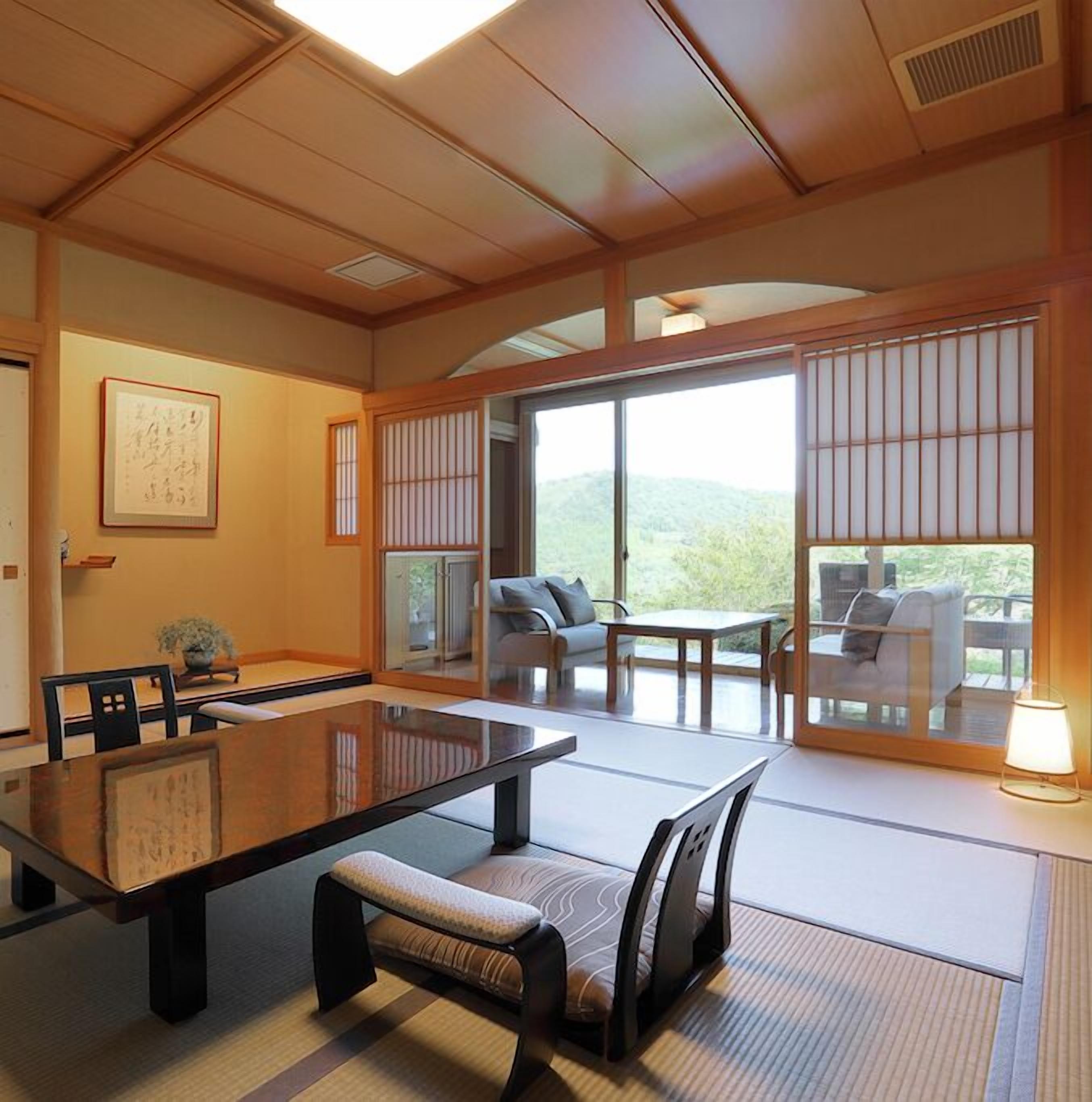 A traditional Japanese-style room with tatami flooring, a low table, and sliding doors leading to a balcony with a view of the hills.