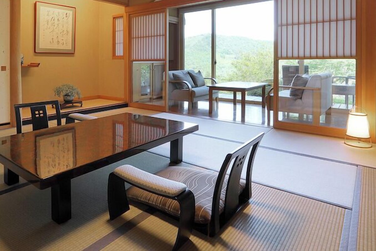 A traditional Japanese-style room with tatami flooring, a low table with cushions, sliding doors, and a view of greenery outside.