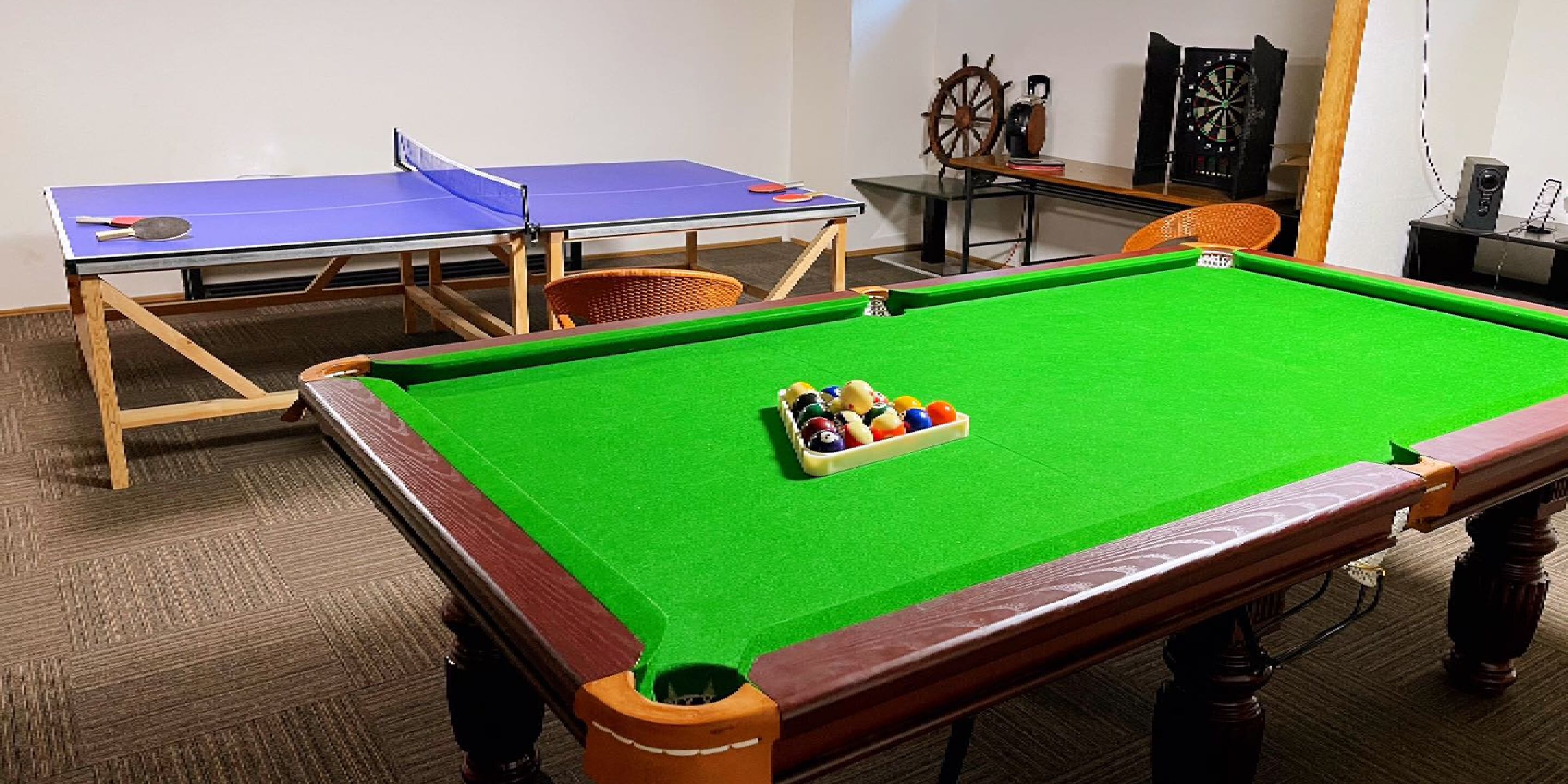 A pool table with balls, a ping pong table, and a dartboard in a room with a carpeted floor.