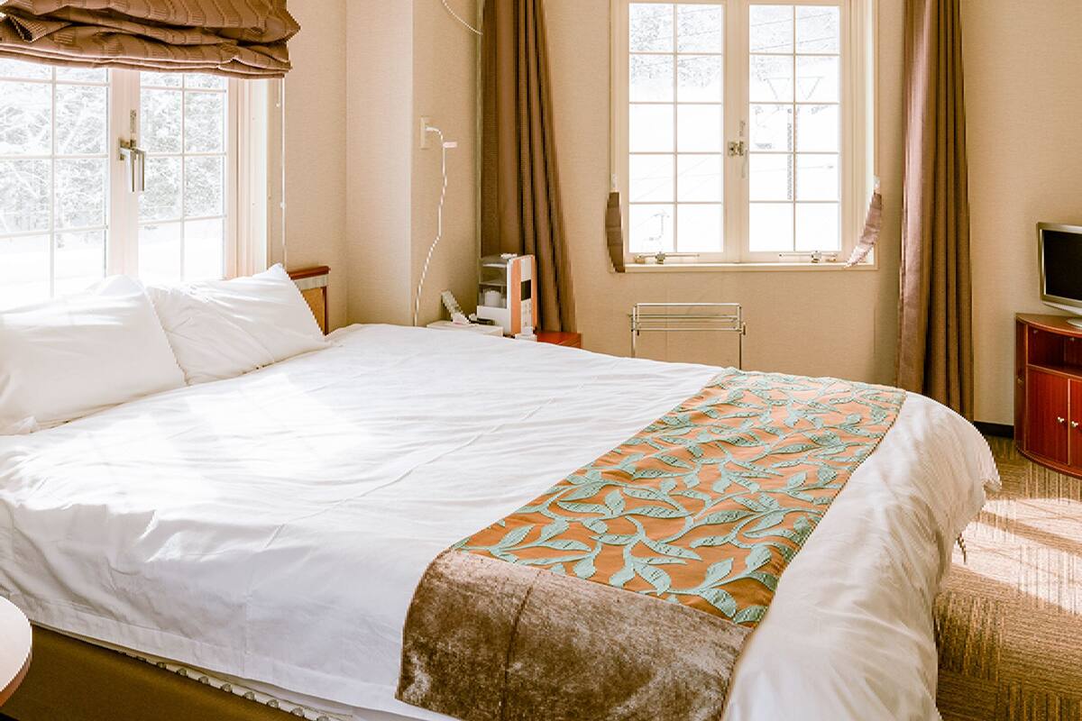 Une chambre d’hôtel avec un grand lit, une table de chevet, un téléviseur posé sur un meuble en bois et une fenêtre avec des rideaux.