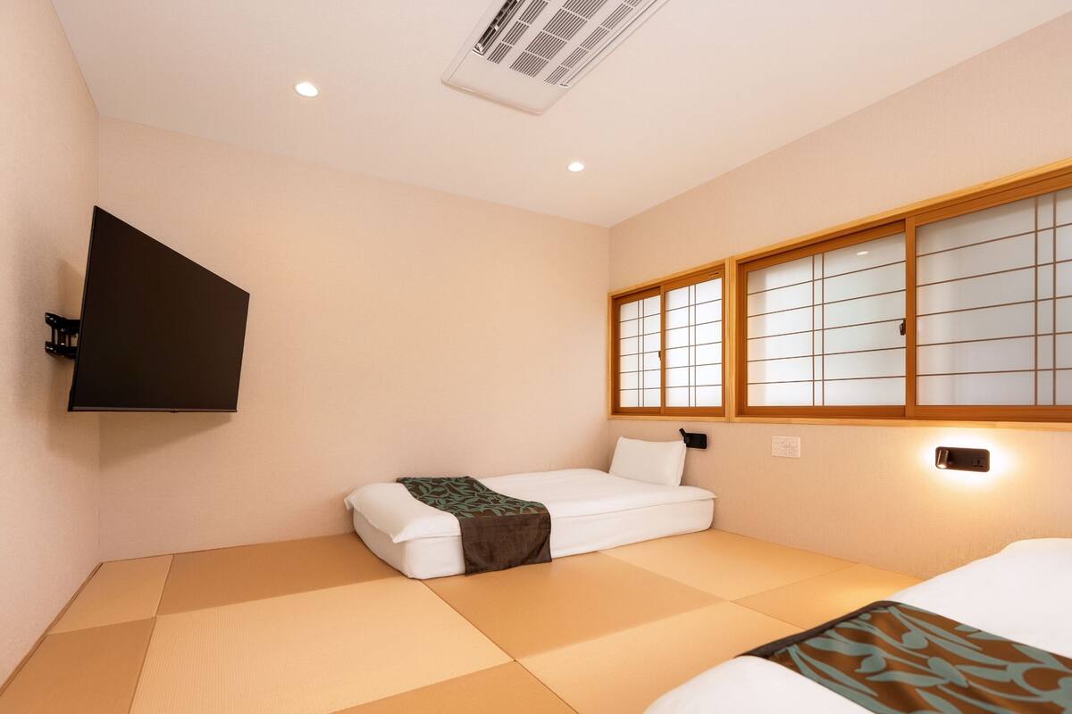Une pièce avec un sol recouvert de tatamis, un lit avec une couvre-lit blanche, un téléviseur fixé au mur et une fenêtre avec des cadres en bois.