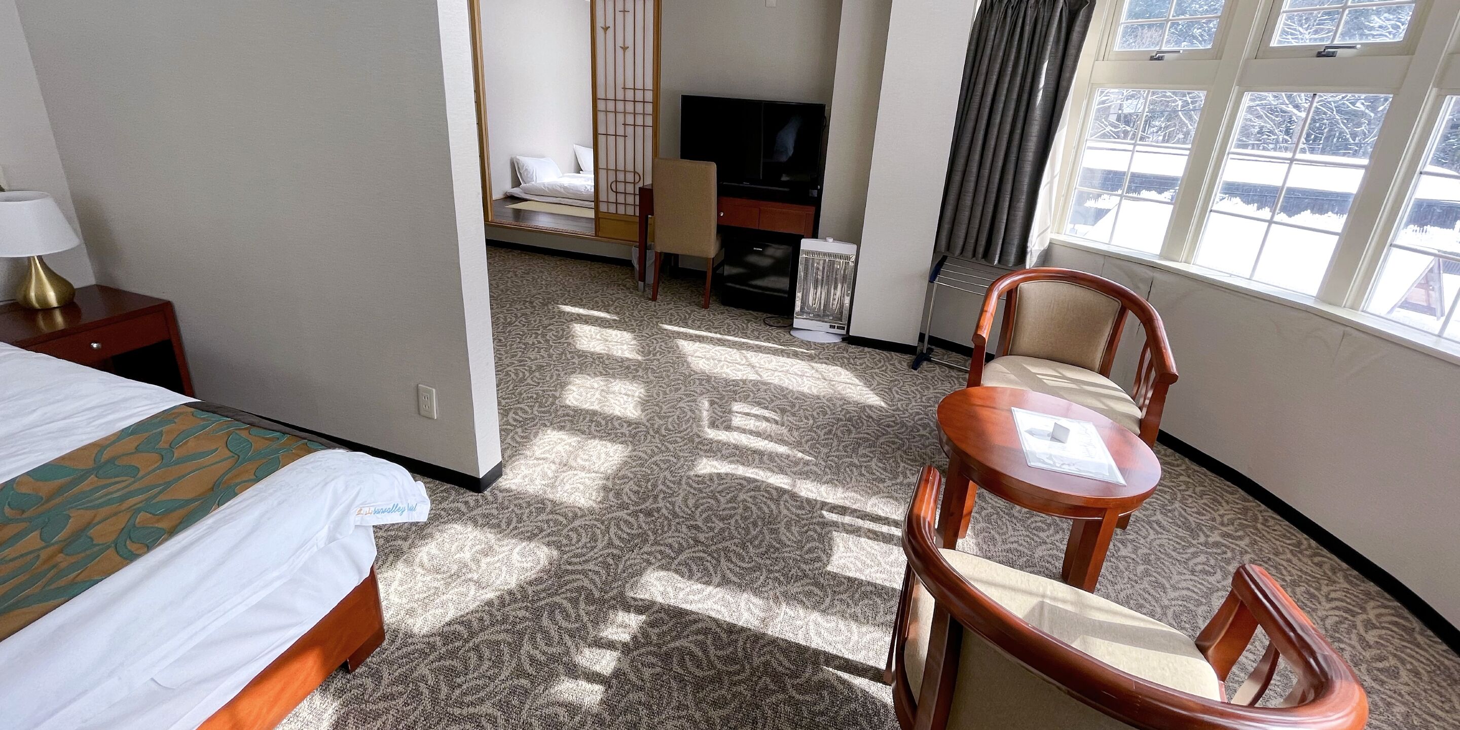 A hotel room with a bed, a desk, a chair, a TV, and a window with a view of a snowy landscape.